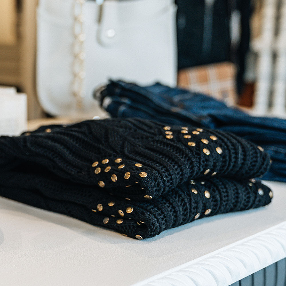 Black knit women's sweater with gold stud accents sitting on a white table top with dark stained distressed jeans and a white handbag
